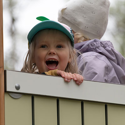 Ein Kleinkind blickt von einem Spielgerät auf dem Spielplatz und lächelt breit.